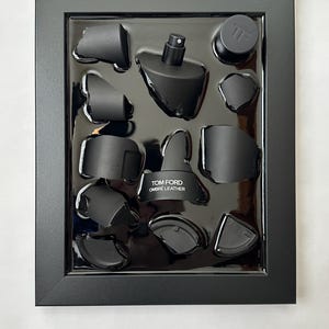 May include: A black framed display featuring a Tom Ford Ombre Leather fragrance bottle and components. The items are arranged in a glossy black tray. The bottle has a black sprayer and the text "TOM FORD OMBRE LEATHER".