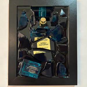 May include: A framed artwork showing a broken blue perfume bottle with gold details. The fragments are arranged in a black frame, with the perfume bottle's label visible, reading "TOM FORD NEROLI PORTOFINO". The artwork is a creative display of shattered glass.