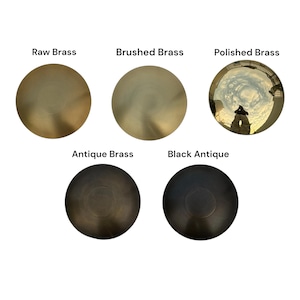 Puede incluir: Cinco discos met&aacute;licos circulares que muestran diferentes acabados: Raw Brass, Brushed Brass, Polished Brass con una superficie reflectante, Antique Brass y Black Antique. Cada disco est&aacute; etiquetado con su nombre de acabado respectivo.