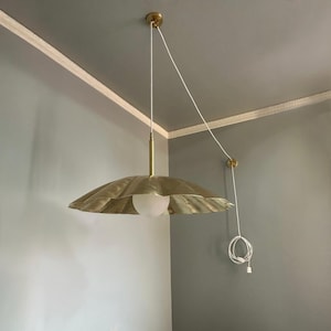 May include: A brass-colored, scalloped pendant light fixture with a white globe bulb. The light hangs from a white cord and has a matching brass ceiling mount. The fixture is suspended in a corner of a room with gray walls.