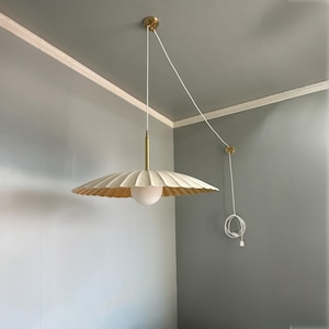 Pode incluir: Uma luminária pendente creme, com bordas onduladas, com interior dourado e uma lâmpada globo branca. A luminária pende de um suporte de teto dourado e um cabo branco. A luminária é um item decorativo para casa.