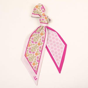 May include: A long, narrow scarf featuring a double-sided design. One side displays a floral pattern with pink and orange flowers on green stems, while the other side has a small pink floral print. The scarf has a bright pink border and the letters AΦ.