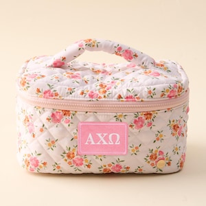May include: A floral print cosmetic bag with a quilted design. The bag features a pink zipper, a handle, and a patch with the Greek letters AXΩ. The bag is decorated with pink, orange, and yellow flowers.