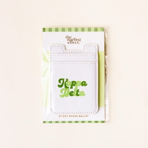 May include: White sticky phone wallet with the text "Kappa Delta" in green lettering. The wallet is packaged on a card with green and white stripes and the words "the darling effect."