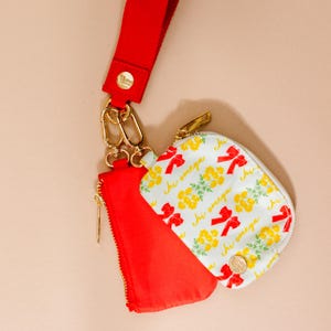 May include: A red lanyard with gold-colored hardware is attached to a red pouch and a white zippered pouch. The white pouch has a floral and bow pattern with the text "Chi Omega".