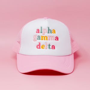 May include: A trucker hat with a light pink brim and mesh back, featuring a white front panel. The text alpha gamma delta is embroidered in the center in a multi-colored font with shades of pink, teal, yellow, and orange thread.