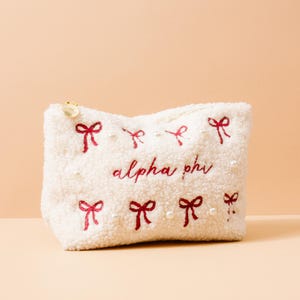 May include: A white, fluffy cosmetic bag with red embroidered bows and the text "alpha phi". The bag has a gold zipper pull and is decorated with small pearl beads. The bag is a soft, textured material.
