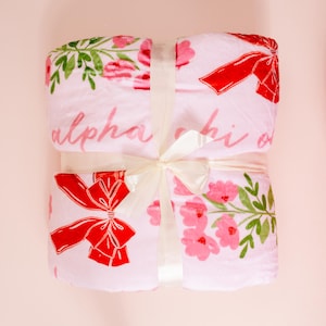 Puede incluir: Una manta rosa doblada con lazos rojos, flores rosas y hojas verdes. La manta está atada con una cinta color crema. Las palabras "alpha chi o" están impresas en rosa.