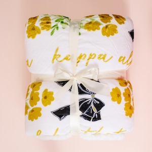 Op de afbeelding: Een witte deken met een bloemenpatroon in geel en groen, en de woorden "kappa al" in gouden letters. De deken is vastgebonden met een crèmekleurig lint en strik. De onderkant van de deken heeft een zwart-wit strik ontwerp.