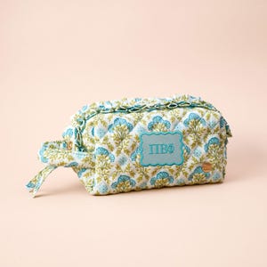 May include: A quilted cosmetic bag featuring a floral pattern in shades of blue and green on a white background. The bag has a ruffled trim, a side handle, and a rectangular teal patch embroidered with the Greek letters Pi Beta Phi in blue.