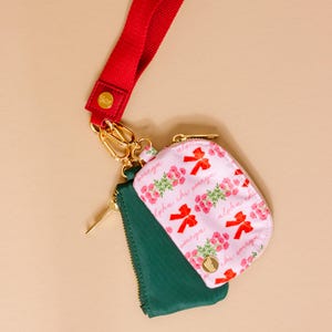 May include: A set of three small zippered pouches. One is pink with a floral and bow pattern, the second is green, and the third is red. The pouches are attached to a red lanyard with gold-colored hardware.