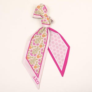 May include: A long, pointed scarf featuring a floral pattern with pink and orange flowers on a light background, bordered by a solid pink trim. One side displays the Greek letters AXΩ in pink. The reverse side has a small, repeating pink floral print.