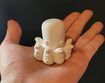 May include: A small, white octopus figurine with a rounded head and eight tentacles. The octopus has detailed suckers on its tentacles and is held in a person's hand. The figurine is made of a smooth, matte material.