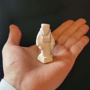 May include: White ceramic figurine of a person with a square head and a long, flowing robe. The figurine is standing on a square base.