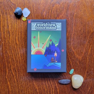 May include: A black box with the text "Worldview Lenormand" in white and a colorful illustration of a sunset, mountains, and a figure with a torch. The box is on a wooden surface.