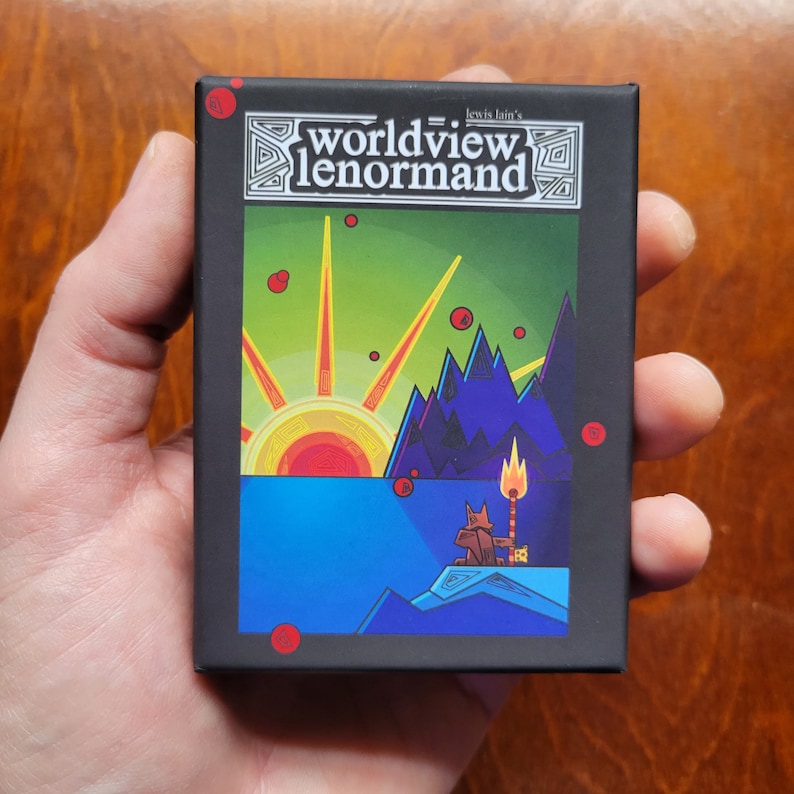 May include: A black box with a colorful illustration of a sunrise over a mountain range with a small animal figure in the foreground. The box has the text "Worldview Lenormand" and "Lewis Iain's" on it.
