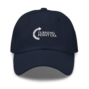 May include: A navy blue baseball cap with a curved brim. The cap features the white text "TURNING POINT USA" and a white arrow logo. The hat is made of a soft material and has a curved brim.