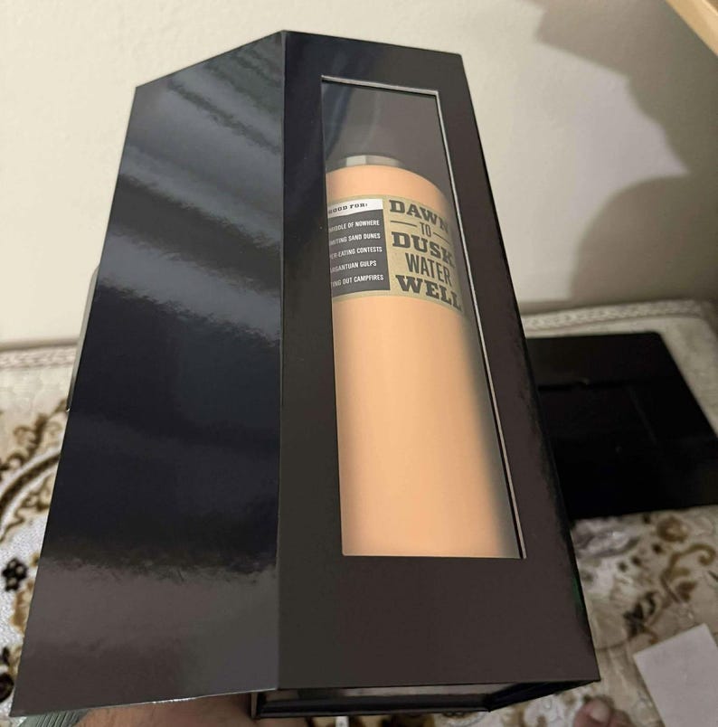 May include: A peach-colored water bottle with the text "Dawn to Dusk Water Well" in a black box with a clear window. The box is black and has a glossy finish. The bottle is cylindrical and appears to be made of metal.