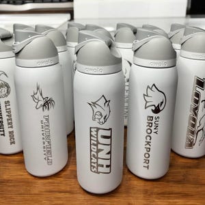May include: A collection of white Owala water bottles with gray lids. Each bottle features a university logo and name in black, including Fairfield University, UNH Wildcats, and SUNY Brockport. The bottles are arranged on a wooden surface.