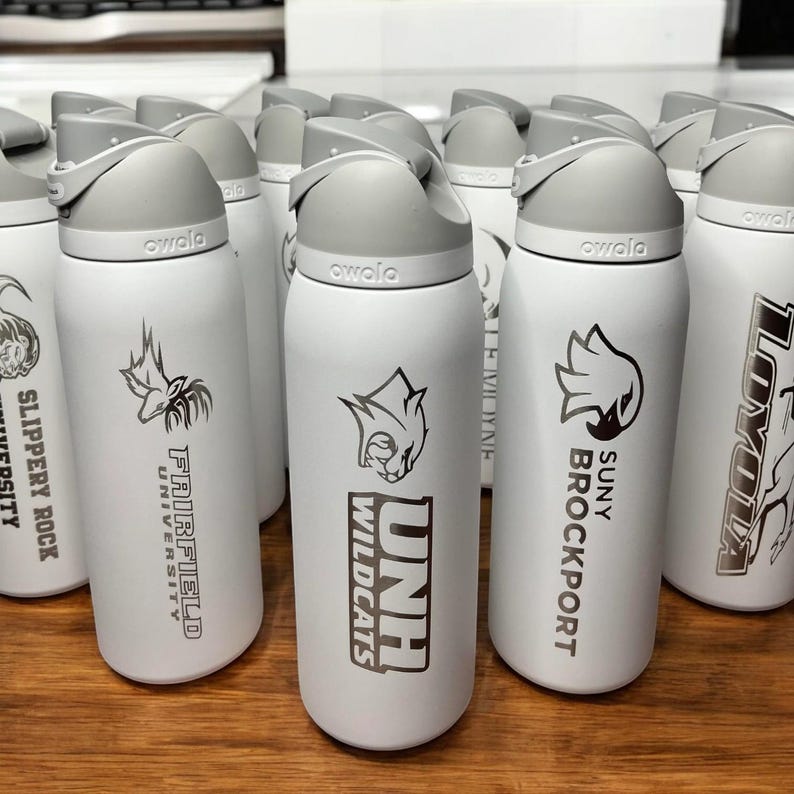 May include: A collection of white Owala water bottles with gray lids. Each bottle features a university logo and name in black, including Fairfield University, UNH Wildcats, and SUNY Brockport. The bottles are arranged on a wooden surface.