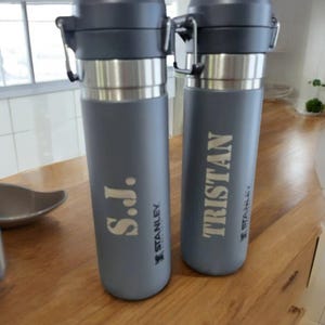 May include: Two grey Stanley water bottles with silver detailing. One bottle has the initials "S.J." and the other has the name "TRISTAN" printed in a light colour. Both bottles have a black cap and a silver clasp.
