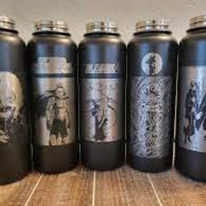Pode incluir: Cinco garrafas de &aacute;gua pretas em a&ccedil;o inoxid&aacute;vel com tampas prateadas e desenhos de personagens de anime gravados. As garrafas s&atilde;o cil&iacute;ndricas e est&atilde;o sobre uma superf&iacute;cie de madeira. Cada garrafa apresenta uma ilustra&ccedil;&atilde;o de personagem diferente.