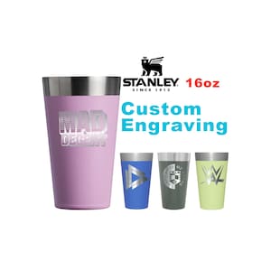 May include: A 16oz Stanley tumbler with custom engraving. The large lavender tumbler features the text MAD DECENT. Three smaller tumblers are shown in blue, grey, and light green, each with a unique silver engraved logo or design on the front.