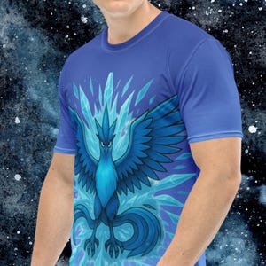 K&ouml;nnte beinhalten: Blaues T-Shirt mit einer stilisierten Illustration eines blauen Vogels mit ausgebreiteten Fl&uuml;geln und Eiskristallen. Der Vogel hat einen grimmigen Ausdruck. Das Shirt ist einfarbig blau und hat kurze &Auml;rmel.