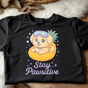 May include: Black t-shirt featuring a cartoon puppy wearing goggles and floating on a pineapple float. The text "Stay Pawsitive" is written in a whimsical font below the puppy. The design includes stars and bubbles.