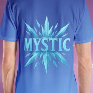 K&ouml;nnte beinhalten: Blaues T-Shirt mit Farbverlauf. Auf der R&uuml;ckseite des Shirts steht das Wort "MYSTIC" in Hellblau, umgeben von einem stilisierten, eisigen Kristall-Design. Das Shirt hat kurze &Auml;rmel.