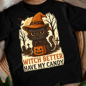 Op de afbeelding: Zwart t-shirt met een cartoon zwarte kat met een heksenhoed en een pompoentas. De tekst "WITCH BETTER HAVE MY CANDY" staat eronder. Het ontwerp bevat bomen, een maan en vleermuizen.