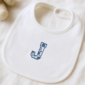 May include: A white baby bib with a blue floral embroidered letter "J" in the center. The bib has a rounded shape with a neck opening at the top. The embroidery features a floral design.