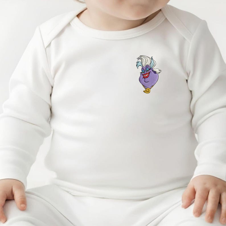 May include: White baby bodysuit with long sleeves. The bodysuit features a colorful cartoon image of Ursula from The Little Mermaid on the front. Ursula is depicted with purple skin, white hair, and a gold necklace.