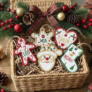 May include: A wicker basket containing four embroidered fabric Christmas ornaments: a gingerbread person labelled Leo, a snowflake labelled Mia, a heart labelled Chloe, and a stocking labelled Noah, plus a Santa face ornament, all with festive decor.