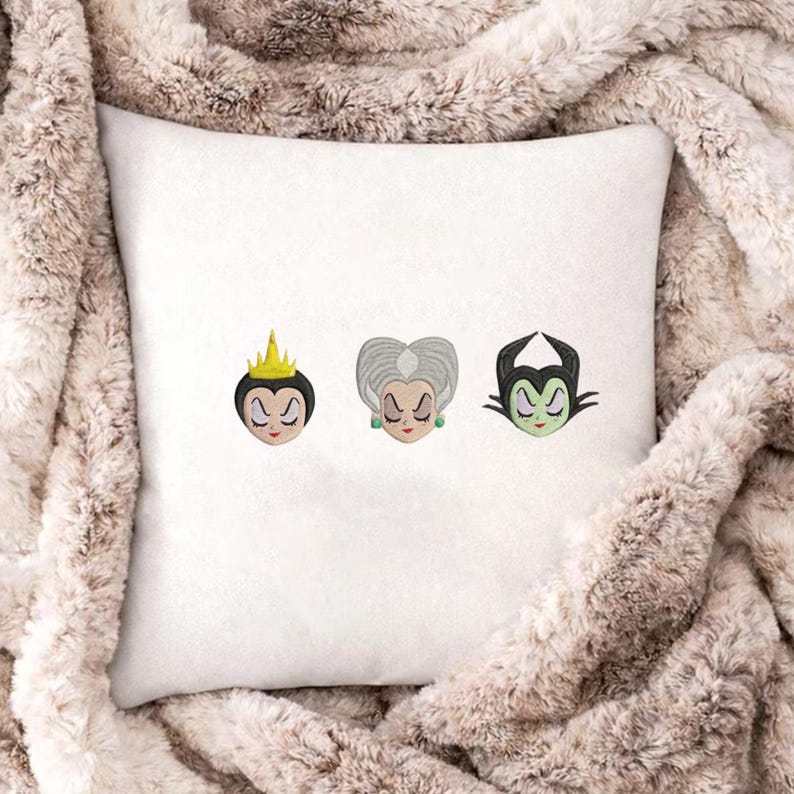 May include: A white square throw pillow featuring three embroidered character faces. The first is a figure with a yellow crown, the second has grey hair and green earrings, and the third is a green-skinned figure with black horns.