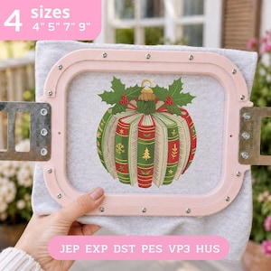 May include: An embroidery design of a Christmas ornament shaped like a pumpkin made of books, featuring green and red spines with gold accents, topped with holly leaves. The design is stitched on light gray fabric held in a light pink embroidery hoop.