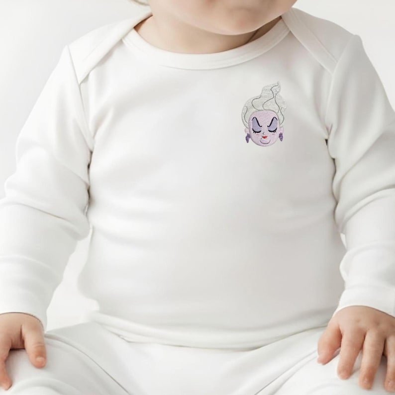 May include: A long-sleeved white baby bodysuit featuring a small embroidered graphic on the upper left chest. The graphic depicts the face of a character with pale skin, white hair, purple earrings, and a stern expression with closed eyes.
