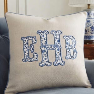 Puede incluir: Un cojín cuadrado de color beige con un monograma grande y centrado con las letras EHB. Las letras están aplicadas en un patrón floral azul y blanco con un borde cosido azul, a juego con el ribete de los bordes del cojín.