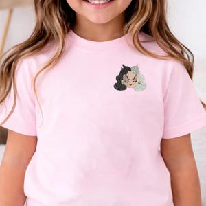 May include: A light pink t-shirt featuring a small embroidered graphic on the upper left chest. The embroidery depicts a stylized face with split black and white hair, closed eyes, red lips, and small green circular earrings on a light background.