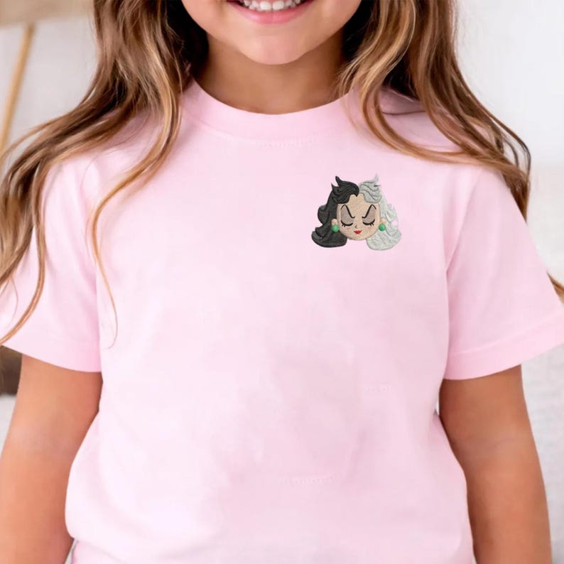 May include: A light pink t-shirt featuring a small embroidered graphic on the upper left chest. The embroidery depicts a stylized face with split black and white hair, closed eyes, red lips, and small green circular earrings on a light background.