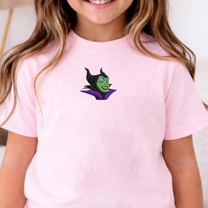 May include: A light pink t-shirt featuring an embroidered image of a green-skinned character with black horns and a purple collar. The character has a sinister smile. The t-shirt is made of soft material.