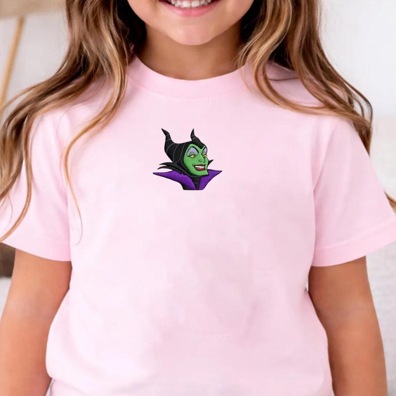 May include: A light pink t-shirt featuring an embroidered image of a green-skinned character with black horns and a purple collar. The character has a sinister smile. The t-shirt is made of soft material.