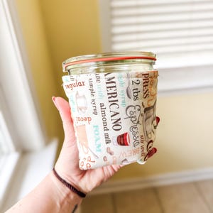 May include: A clear glass jar with a red-rimmed lid, wrapped in a fabric coffee sleeve. The sleeve features coffee-themed text and graphics, including "AMERICANO" and "Espresso". The jar is held in a person's hand.