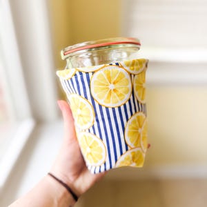 May include: A clear glass jar with a red-rimmed lid is wrapped in a fabric cozy. The cozy features a pattern of yellow lemon slices on a white background with blue vertical stripes. The jar is held in front of a blurred background.