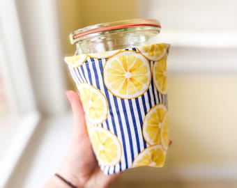May include: A clear glass jar with a red-rimmed lid is wrapped in a fabric cozy. The cozy features a pattern of yellow lemon slices on a white background with blue vertical stripes. The jar is held in front of a blurred background.