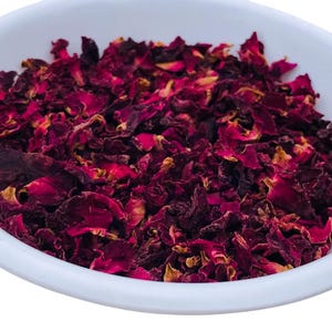 May include: A white, oval-shaped bowl filled with dried rose petals. The petals are a deep burgundy color with hints of yellow and brown. The image is a close-up shot, highlighting the texture and color of the petals.