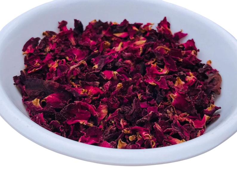 May include: A white, oval-shaped bowl filled with dried rose petals. The petals are a deep burgundy color with hints of yellow and brown. The image is a close-up shot, highlighting the texture and color of the petals.