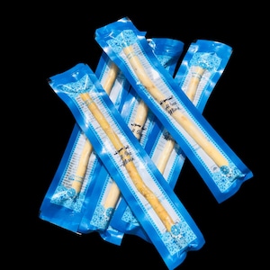 May include: Several miswak sticks, a natural teeth-cleaning tool, are displayed. Each stick is individually packaged in a blue and white plastic wrapper. The wrappers have a decorative pattern and text in a foreign language. The sticks are a light beige color.