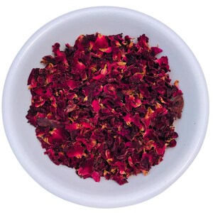 May include: A white ceramic bowl filled with dried red rose petals. The petals are a deep burgundy color with some lighter, yellowish hues. The bowl is round and sits on a white background. The image is a close-up shot.