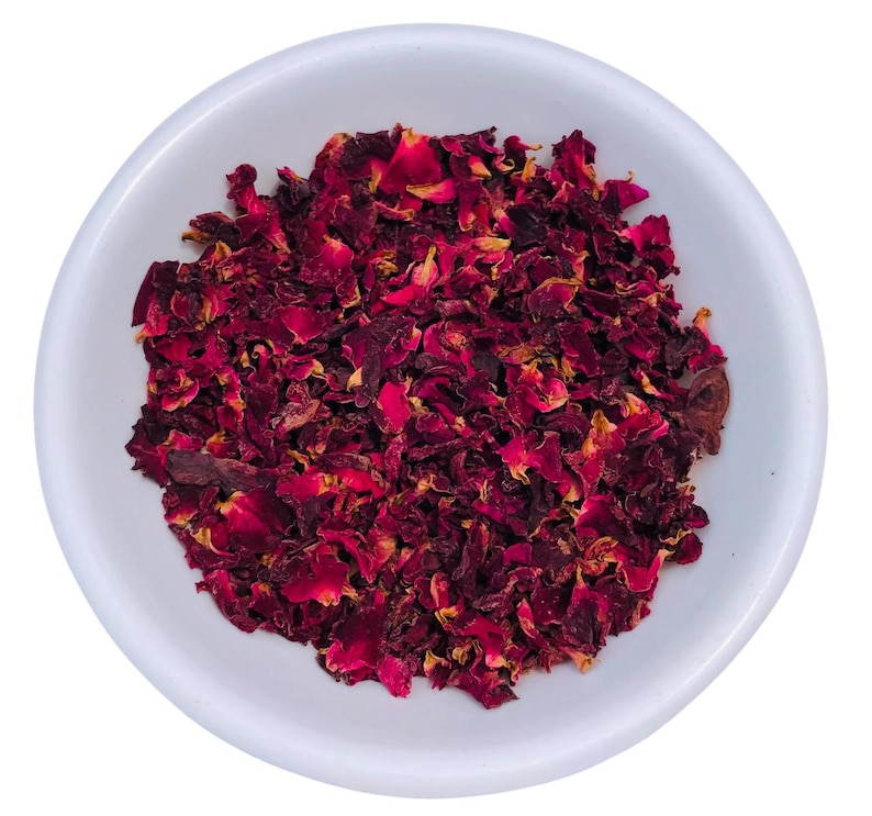 May include: A white ceramic bowl filled with dried red rose petals. The petals are a deep burgundy color with some lighter, yellowish hues. The bowl is round and sits on a white background. The image is a close-up shot.